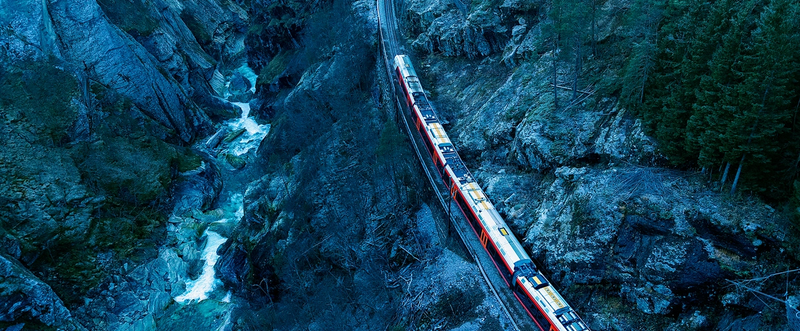 Foto: Et tog kjører snart inn i en tunnel i et svært bratt berg. Til venstre i bildet renner en elv i et juv. Jernbanen ligger parallelt med elveleiet, men lenger opp i skrenten. Toget kommer fra nederste høyre kant av bildet og kjører mot tunnelen som ligger lengt opp og i senter av bildet.