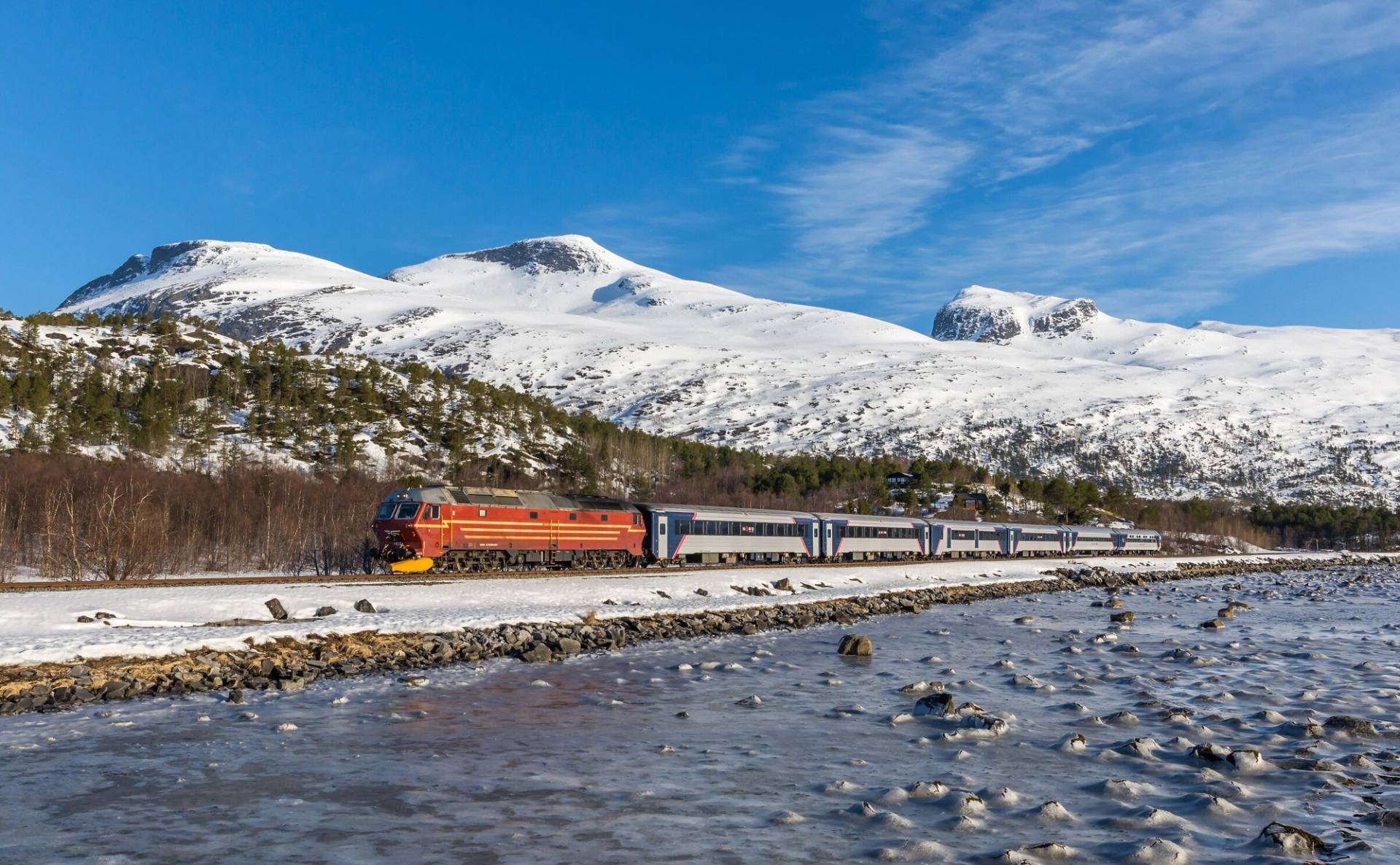 Tog som passerer mellom et islagt vann og snødekte fjell. Blå himmel og spredte skyer.