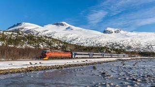 Tog som passerer mellom et islagt vann og snødekte fjell. Blå himmel og spredte skyer.