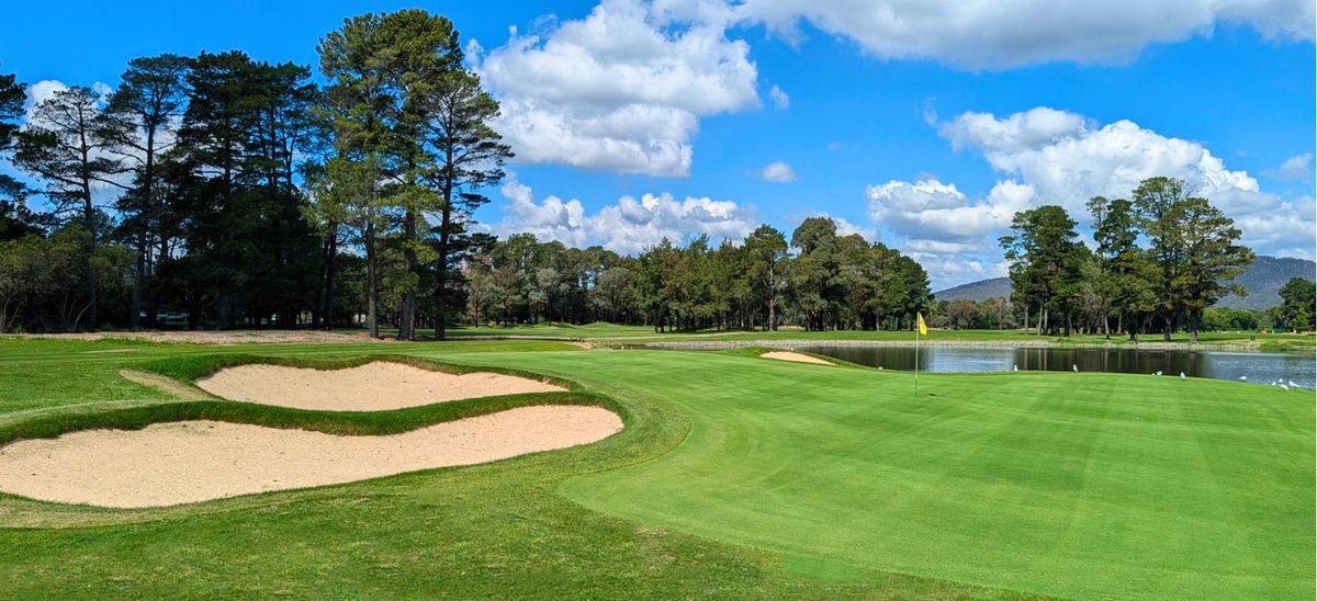 A green with yellow flag and pond at Yowani Country Club, framed by bunkers and tall pine trees.