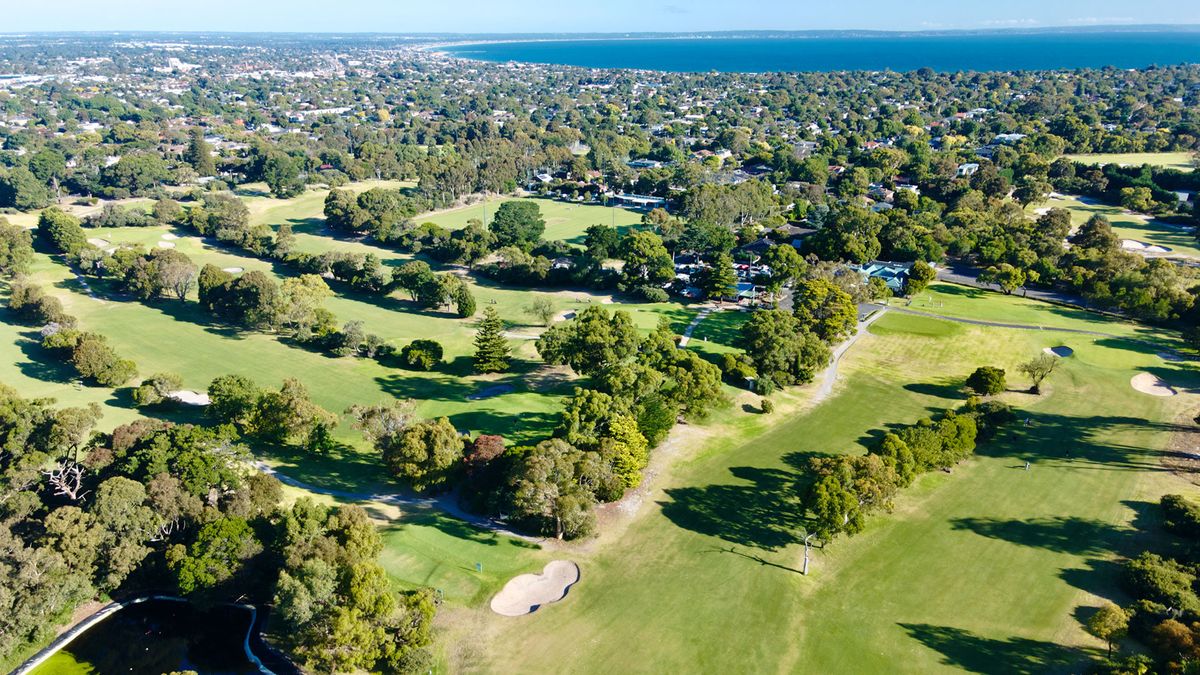 Aerial view of Cheltenham Golf Club's tree-lined fairways and clubhouse, with Port Phillip Bay in the background.