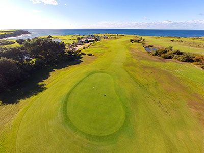 Aerial view of a green at The Coast Golf Club with the ocean, coastal dunes, and clubhouse buildings in the background.