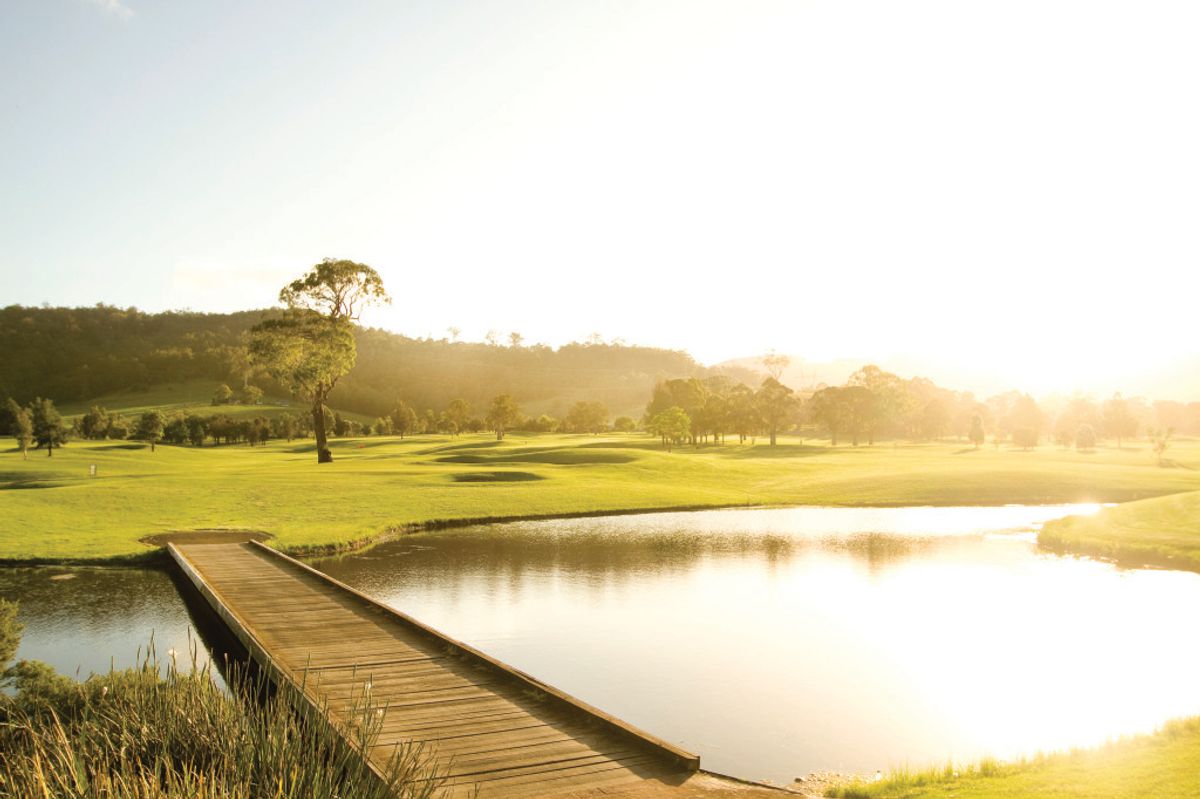 A pond with a wooden footbridge and fairway at Calderwood Valley Golf Course, lit by golden afternoon sun.