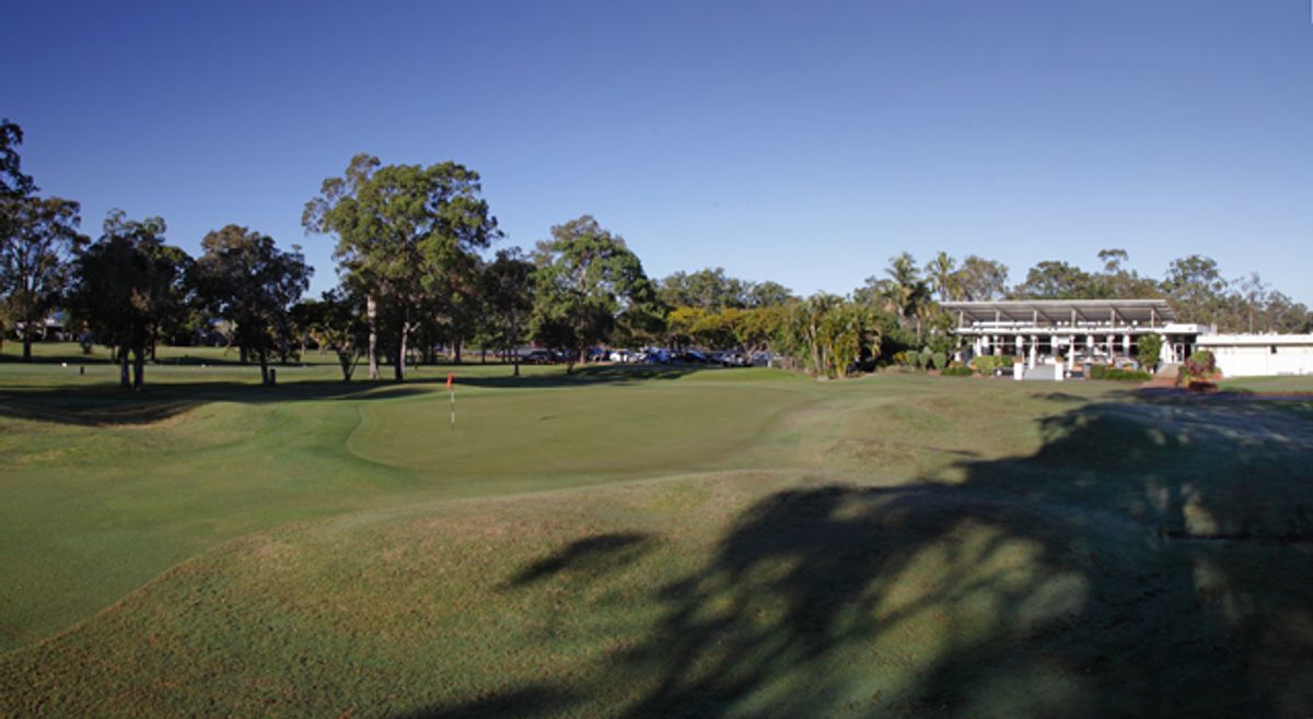 The 18th green at Virginia Golf Club with a red flag, backed by the clubhouse and parked cars among trees.