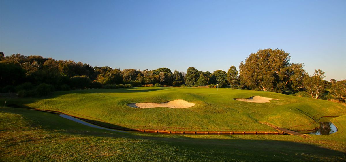 A bunkered green at Eastlake Golf Club lit by golden-hour sun, with a creek crossing in the foreground.