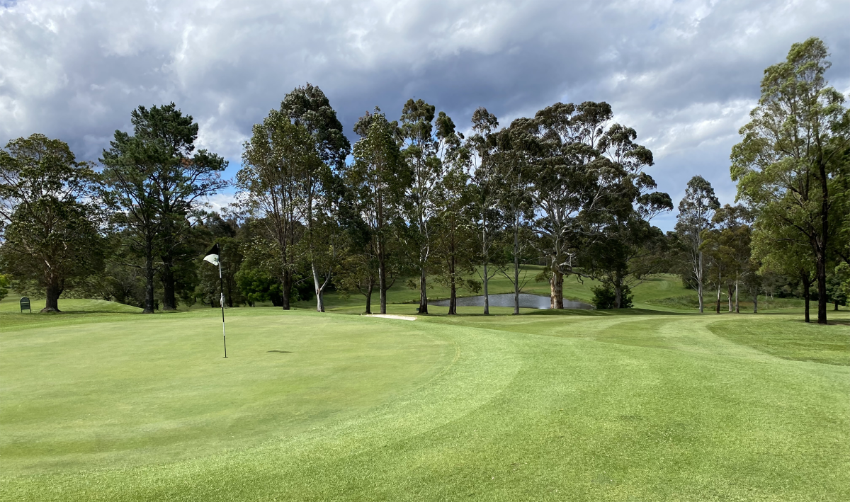 The 9th green at Springwood Country Club with a white flag, sand bunker, small pond, and tall gum trees under a cloudy sky.