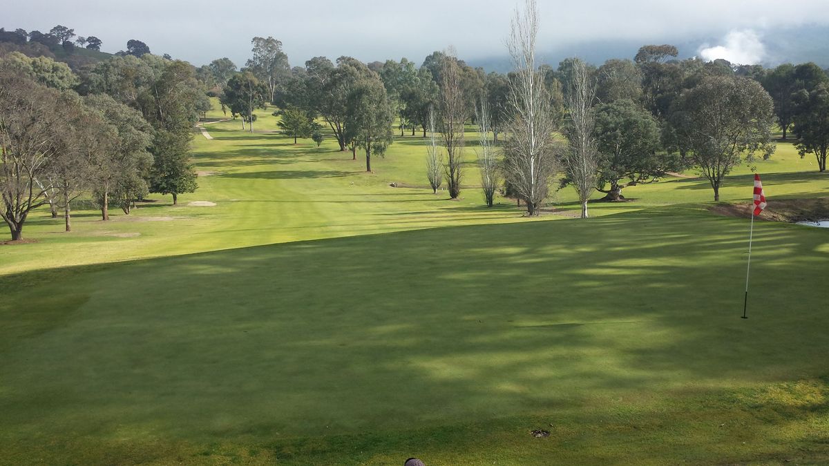 A fairway leading to a green with red and white flag at Tumut Golf Club, lined with trees under a cloudy sky.