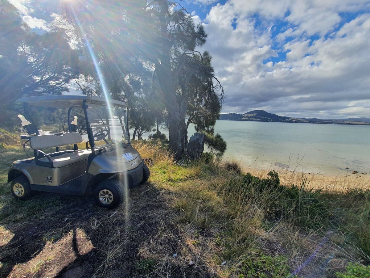 A golf cart parked on a grassy point overlooking the beach and bay at Tasmania Golf Club, with hills visible across the water.