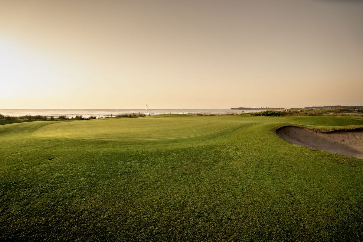 A seaside green with bunker at Belmont Golf & Bowls, overlooking the ocean at sunset.