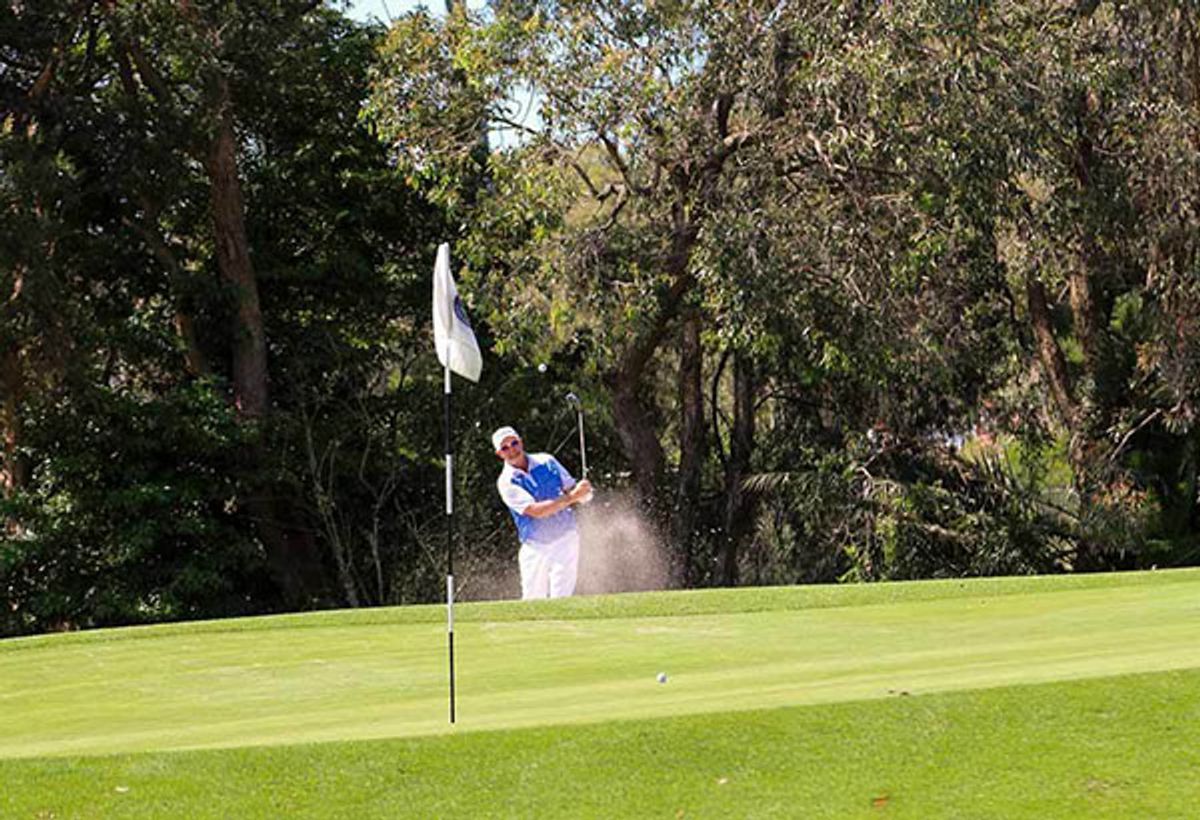 A golfer playing a bunker shot onto the green at Waratah Golf Club, with a white flag and trees in the background.