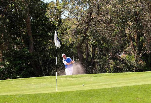 A golfer playing a bunker shot onto the green at Waratah Golf Club, with a white flag and trees in the background.