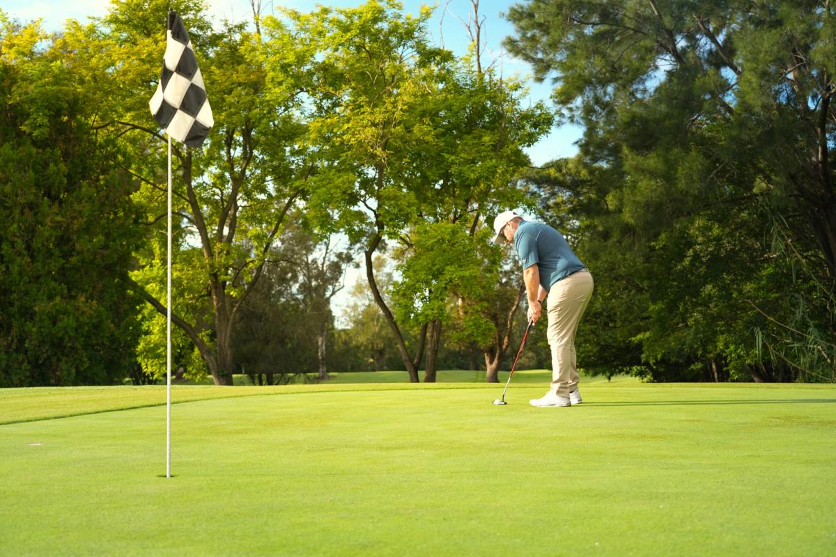 A golfer putting on a green at The Grange Golf Club, with a checkered flag and tall trees surrounding the green.