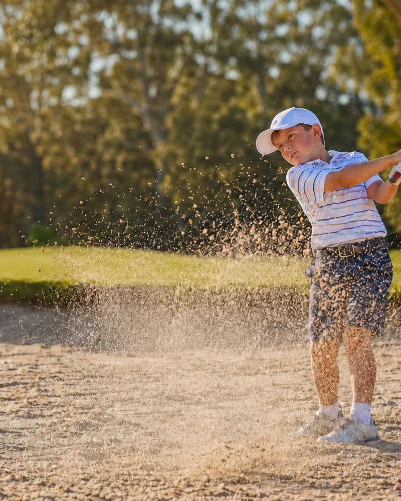 A young golfer in a white cap and striped polo hitting from a sand bunker, sending sand and the ball flying on an Australian golf course