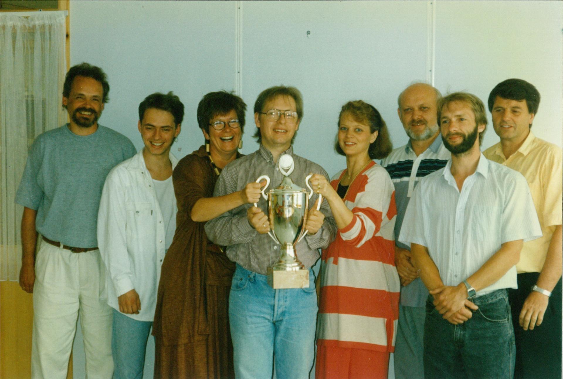 An older photo from 1982 showing seven smiling Solwr employees holding up a trophy