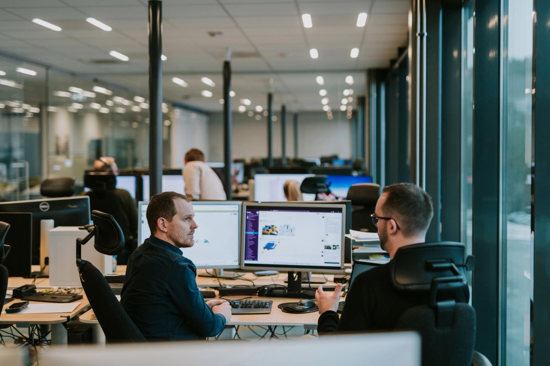 Two male Solwr employees sitting at an office desk talking to each other while pointing at the screen in front of them