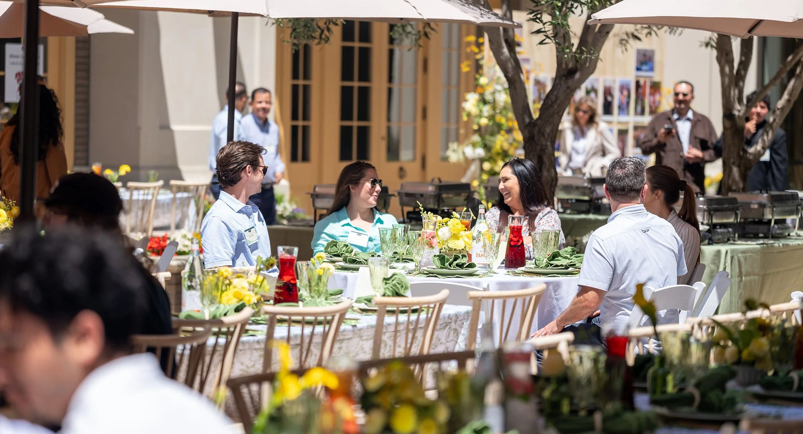 Guests seated at round tables on the Portobello's patio under cream umbrellas, with olive trees, yellow flowers and folded green napkins on the linen.