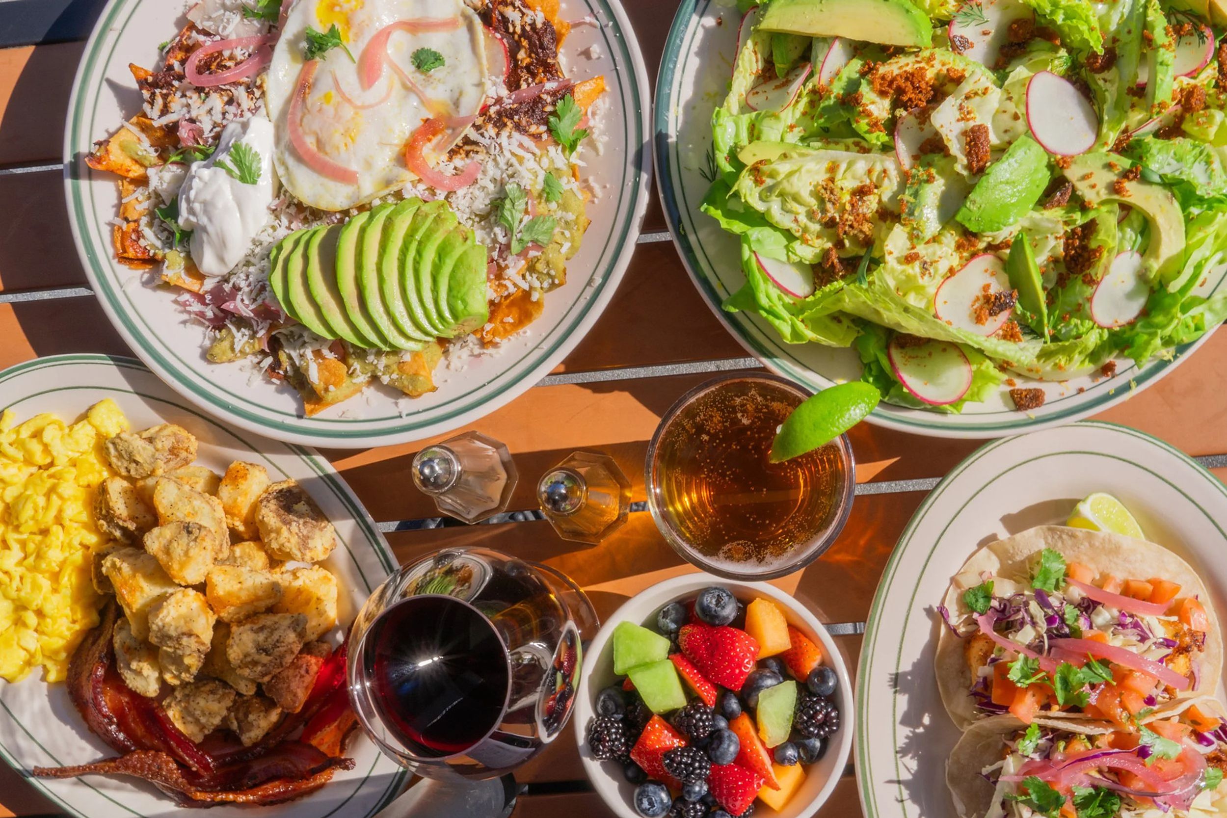 An overhead table on the patio: chilaquiles under a fried egg with sliced avocado, a gem lettuce salad with radish, scrambled eggs with bacon and potatoes, fish tacos, a bowl of berries and melon, a glass of red wine and a beer.