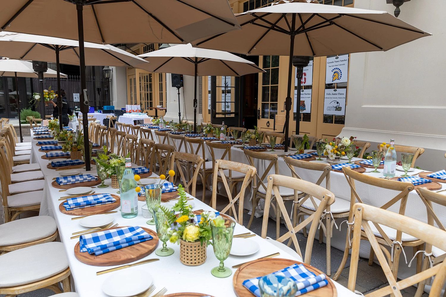 Guests seated along a laid table on the patio, blue gingham napkins on wooden boards, jars of yellow flowers down the middle.