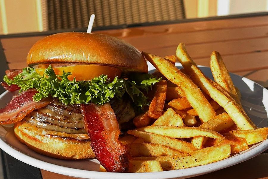 A bacon burger on a brioche bun with lettuce and tomato, piled beside seasoned steak fries on a white plate, on a wooden patio table.