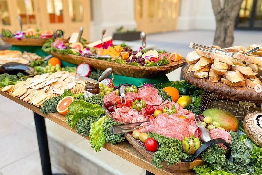 A grazing table set outdoors: wooden boards of cured meats, cheeses, olives, sliced fruit and baguette, banked with kale and edible flowers.