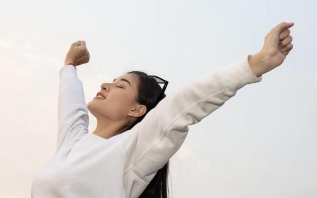 Een jonge vrouw in witte trui strekt blij haar armen omhoog tegen een heldere lucht
