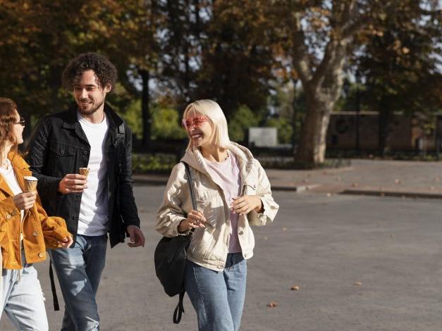 A group of three people dressed in casual autumn attire are joyfully walking on a sunlit street, carrying cups and bags, with autumn trees in the background.