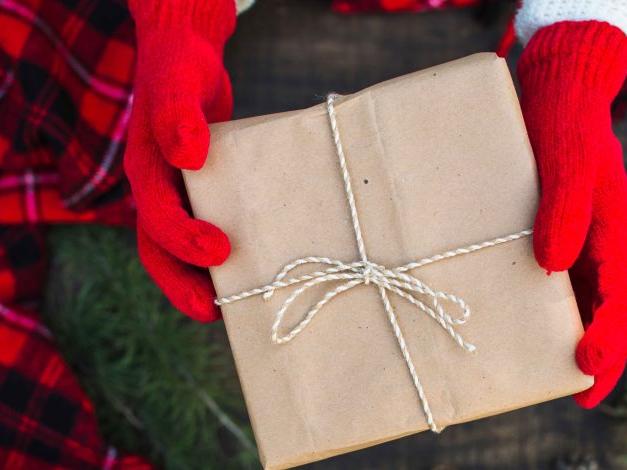 A person wearing bright red gloves holds a neatly wrapped brown paper gift tied with twine, set against a backdrop of festive red plaid fabric and green pine branches, creating a cozy holiday atmosphere.