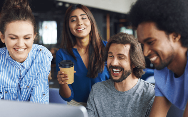 A diverse group of four smiling people gather around a laptop, with one person holding a coffee cup, suggesting a collaborative work environment in a casual office setting.