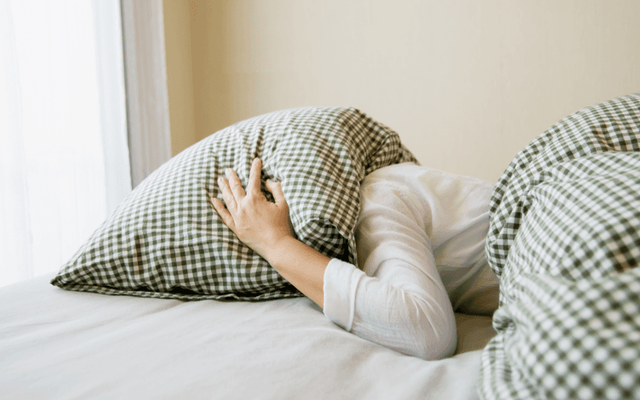 A person covers their head with a checkered pillow while lying in bed, creating a cozy and restful atmosphere in a softly lit room.