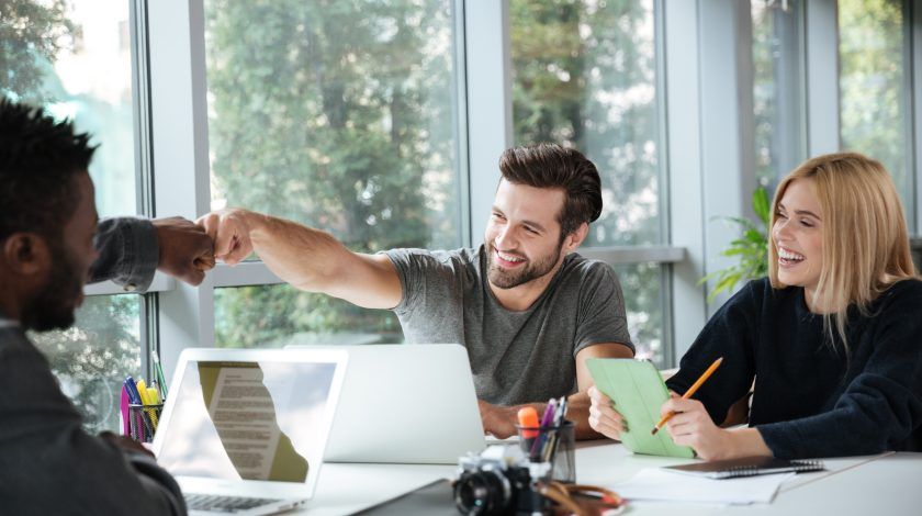A diverse group of people in a bright, modern office setting engage in a cheerful meeting, with one person fist-bumping another across a table cluttered with laptops, notebooks, and a camera.
