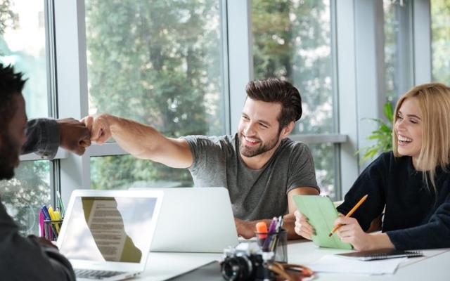 A diverse group of people in a bright, modern office setting engage in a cheerful meeting, with one person fist-bumping another across a table cluttered with laptops, notebooks, and a camera.