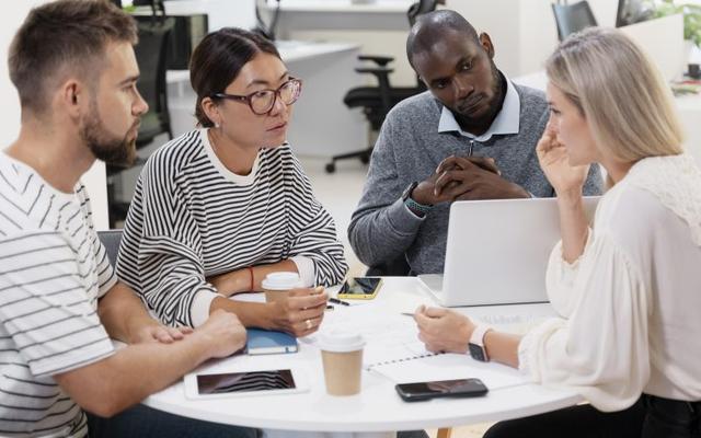 Four diverse colleagues engage in a collaborative discussion around a white table in a modern office, with notebooks, a laptop, and coffee cups visible.