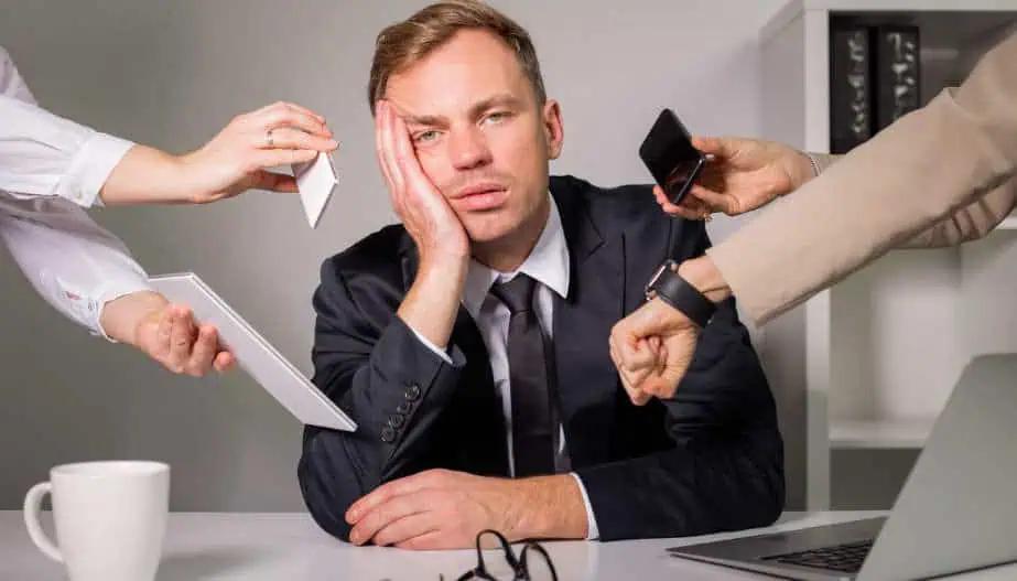 In an office setting, a weary businessman in a suit rests his face on his hand, surrounded by multiple outstretched arms holding a phone, a tablet, a document, and a watch, symbolizing overwhelming demands and stress.