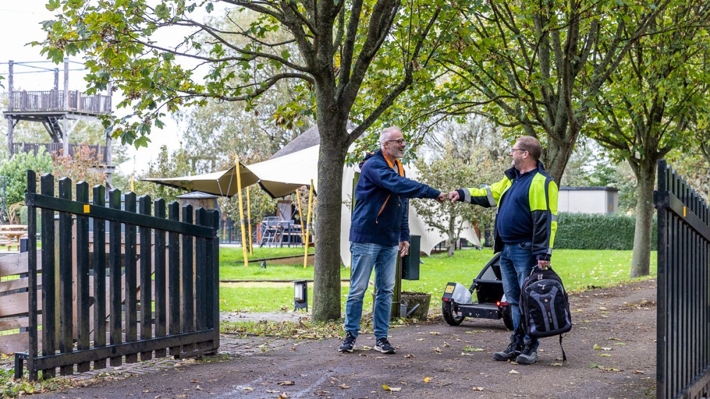 Two people engage in conversation near a stone gate, surrounded by trees and greenery on a pleasant day.
