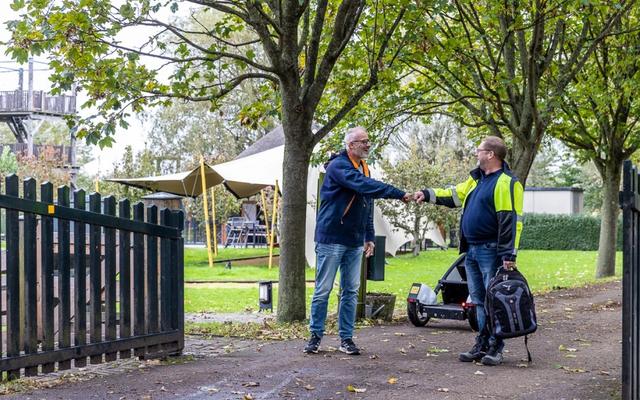 Two people engage in conversation near a stone gate, surrounded by trees and greenery on a pleasant day.