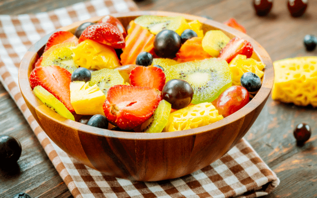 A wooden bowl filled with a vibrant assortment of fresh fruit, including sliced strawberries, kiwis, pineapples, grapes, and blueberries, rests on a checkered cloth atop a wooden table.