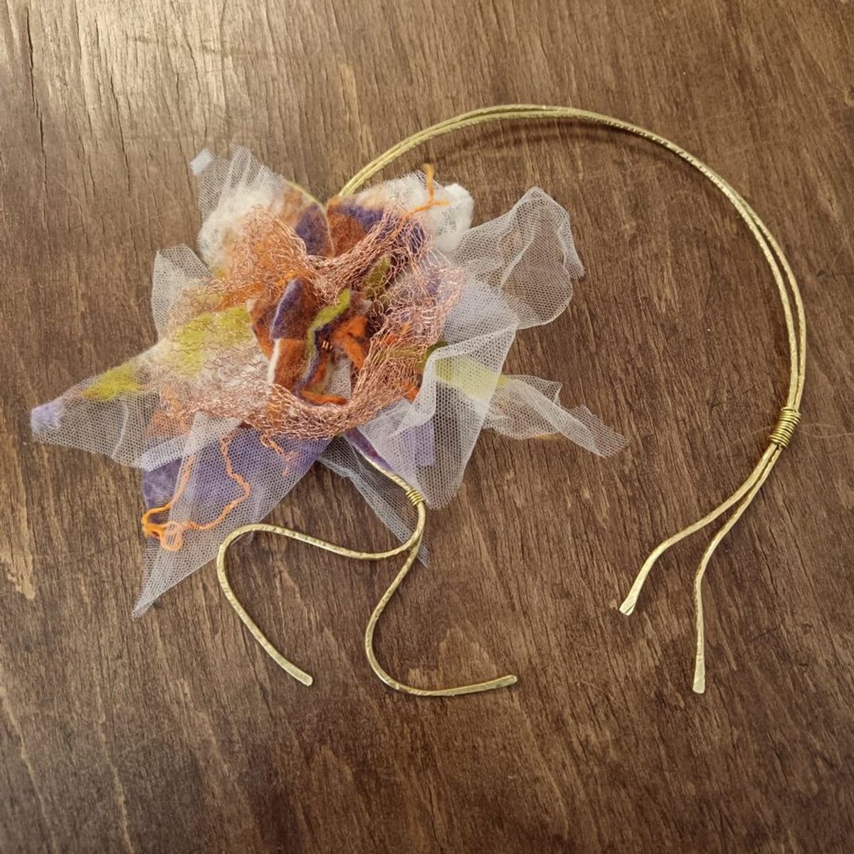 Collier in ottone a due fili con un fiore di feltro viola, arancio e verde, tulle e rete metallica, su un tavolo di legno