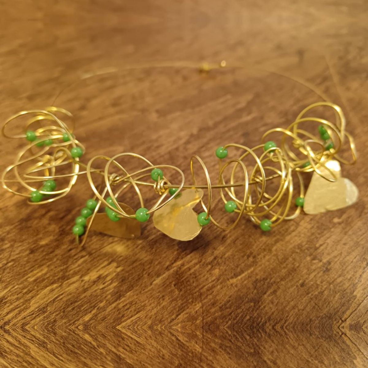Collana in filo di ottone con cerchi intrecciati, cristalli verdi e tre cuori, appoggiata su un tavolo di legno