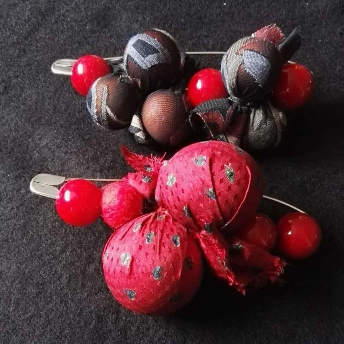 Due spille con palline di stoffa ricavata da cravatte, una rossa e una marrone e grigia, con perle rosse, su un panno scuro