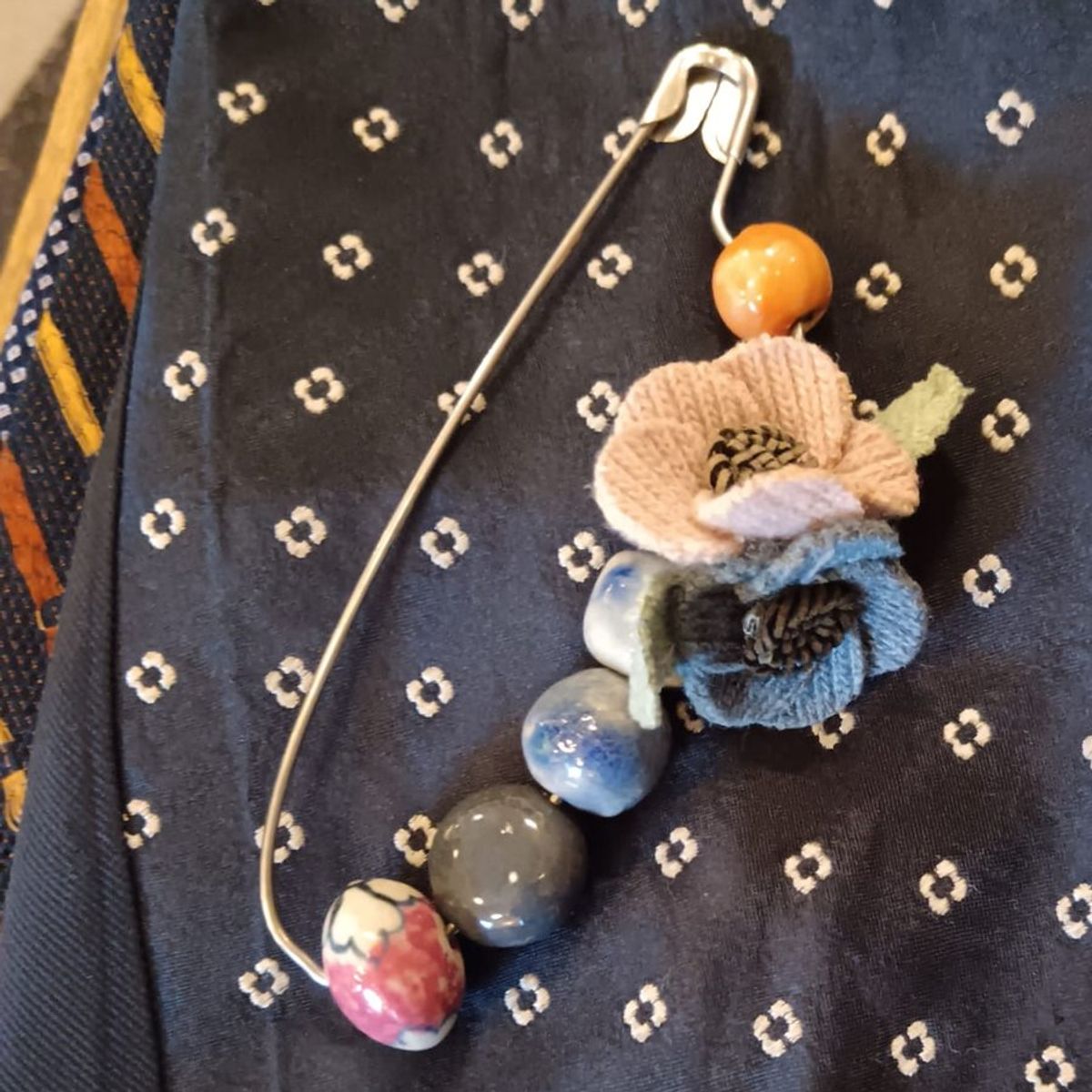 La stessa spilla vista dall'alto: perle di ceramica arancio, blu, grigia e decorata, con i fiori di lana