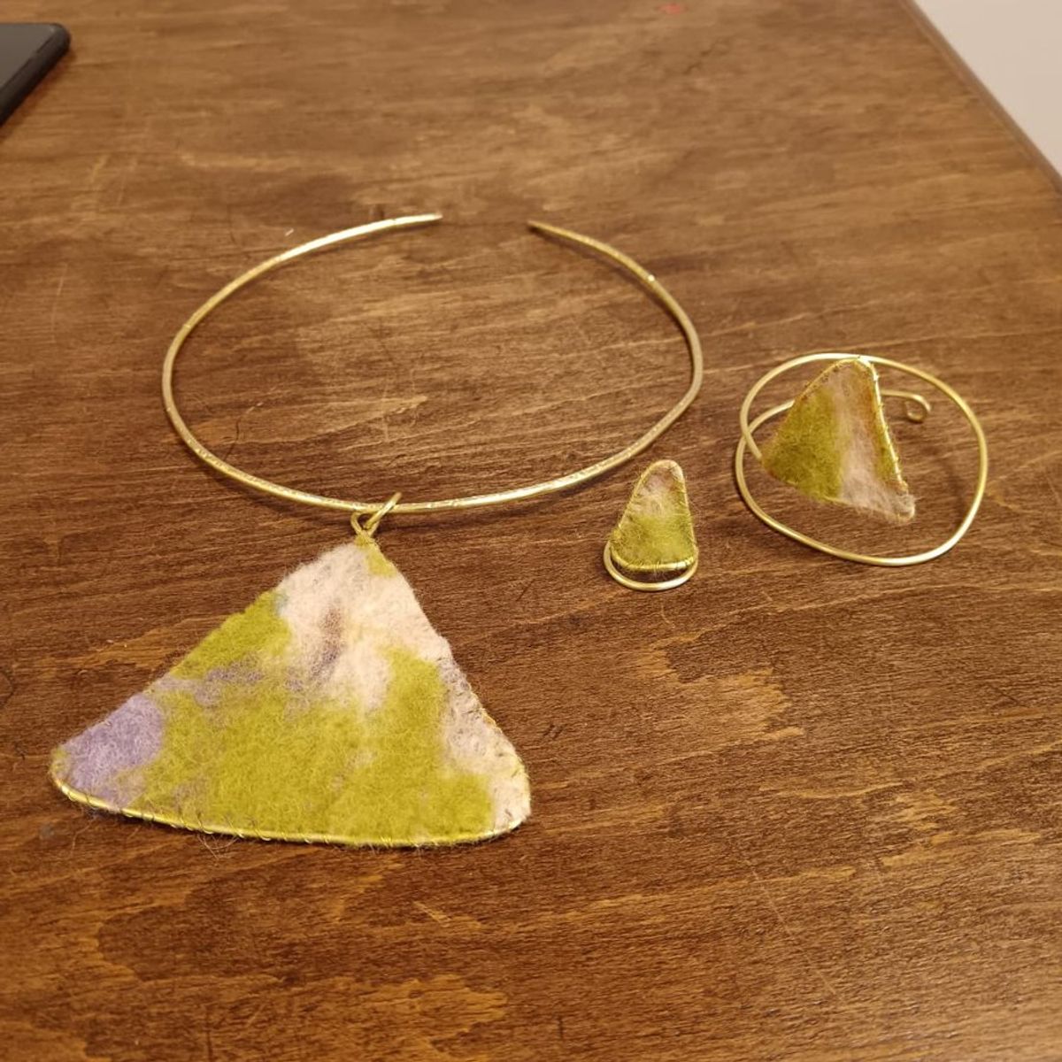 Parure in ottone con triangoli di feltro verde, bianco e lilla: collier con pendente, anello e bracciale su un tavolo di legno