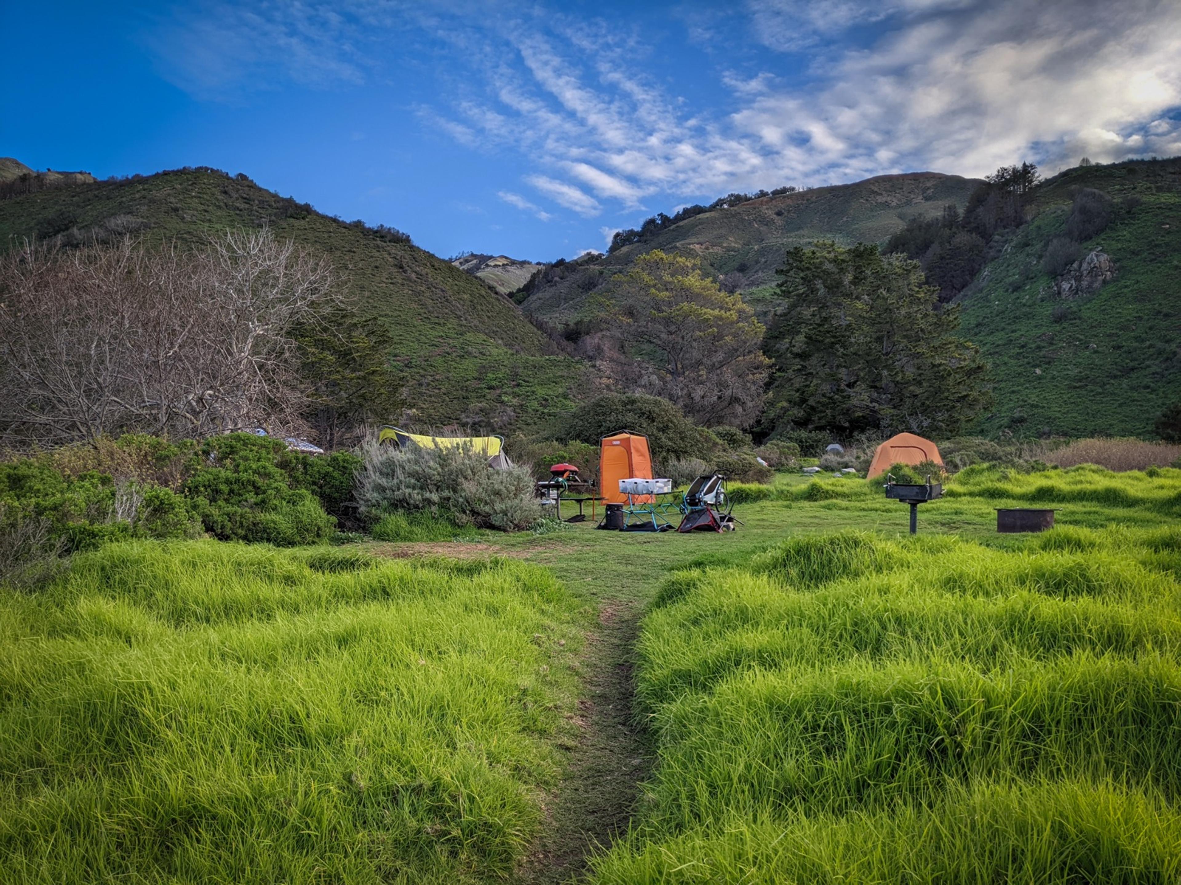 A Kirk Creek campsite with green spring grass, tents, a picnic table behind brush, and a fire ring out in the meadow A Kirk Creek campsite with green spring grass, tents, a picnic table behind brush, and a fire ring out in the meadow