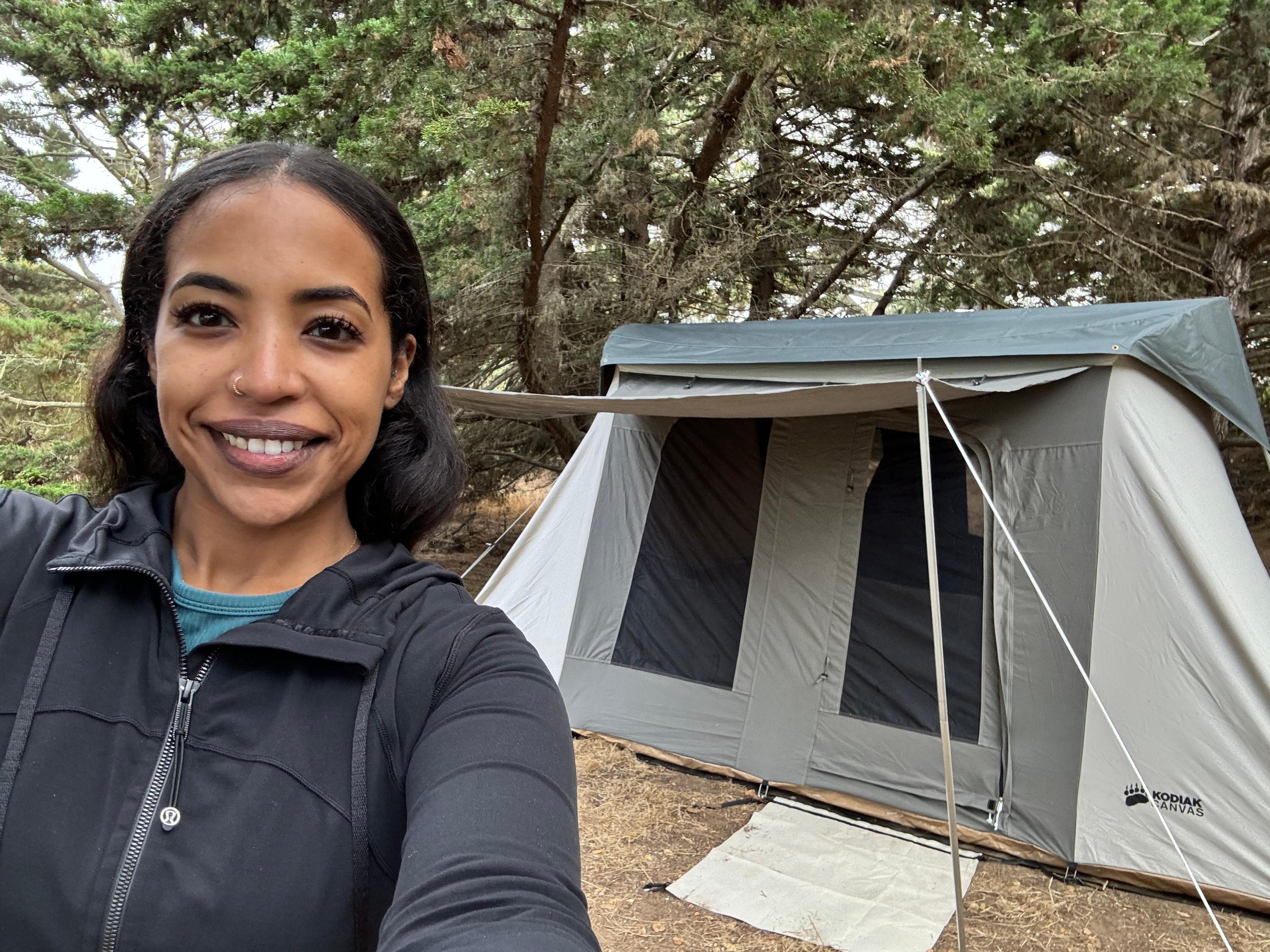 First-time camper Maab proudly posing next to her freshly assembled Kodiak Flex-Bow 10x10 canvas tent at a group trip campsite