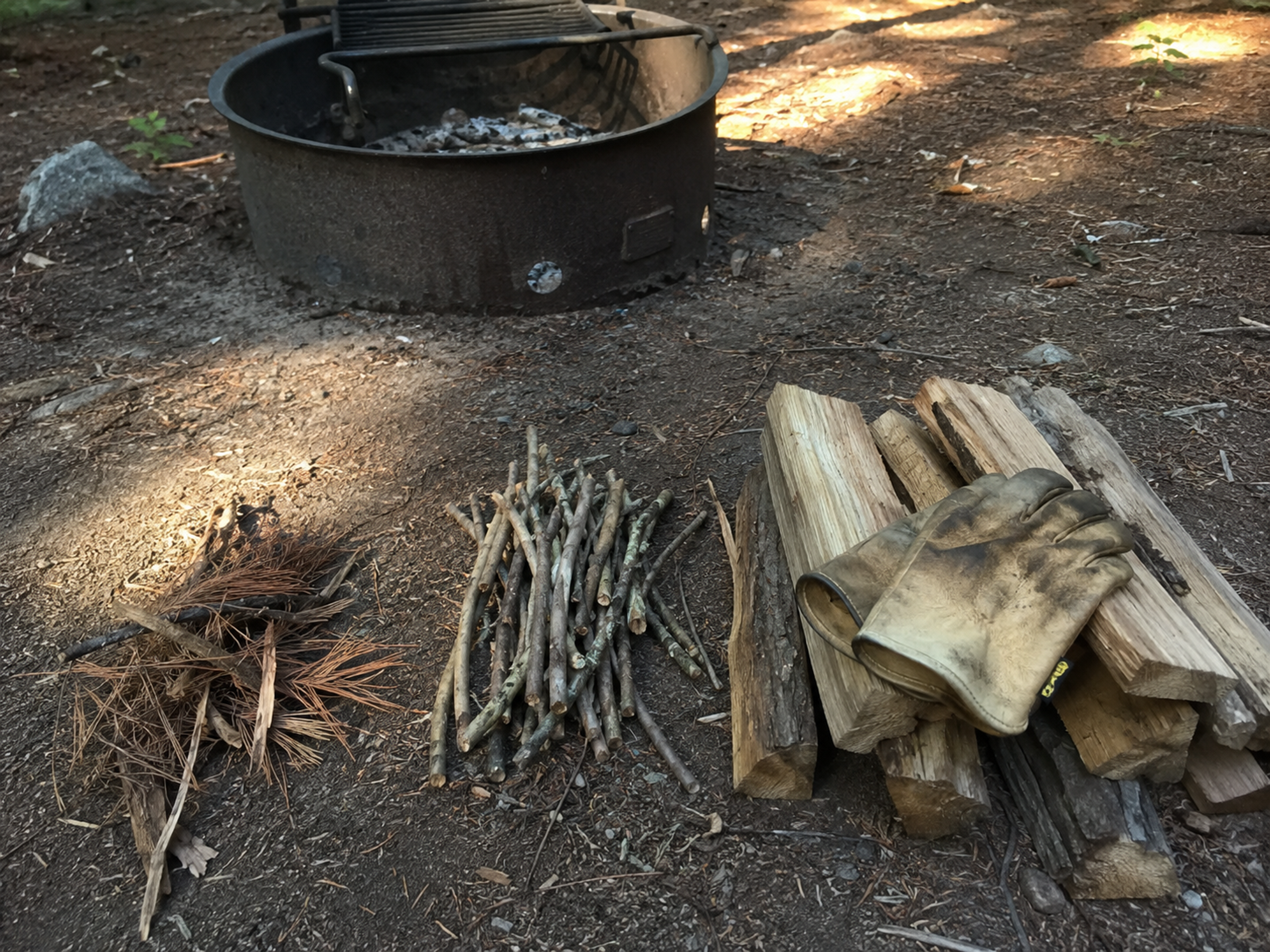 Three sorted piles of wood on dirt next to a metal campground fire ring at golden hour: dry pine needles and bark strips on the left, pencil-thick kindling sticks in the middle, split firewood logs on the right with dirty work gloves resting on top Three sorted piles of wood on dirt next to a metal campground fire ring at golden hour: dry pine needles and bark strips on the left, pencil-thick kindling sticks in the middle, split firewood logs on the right with dirty work gloves resting on top