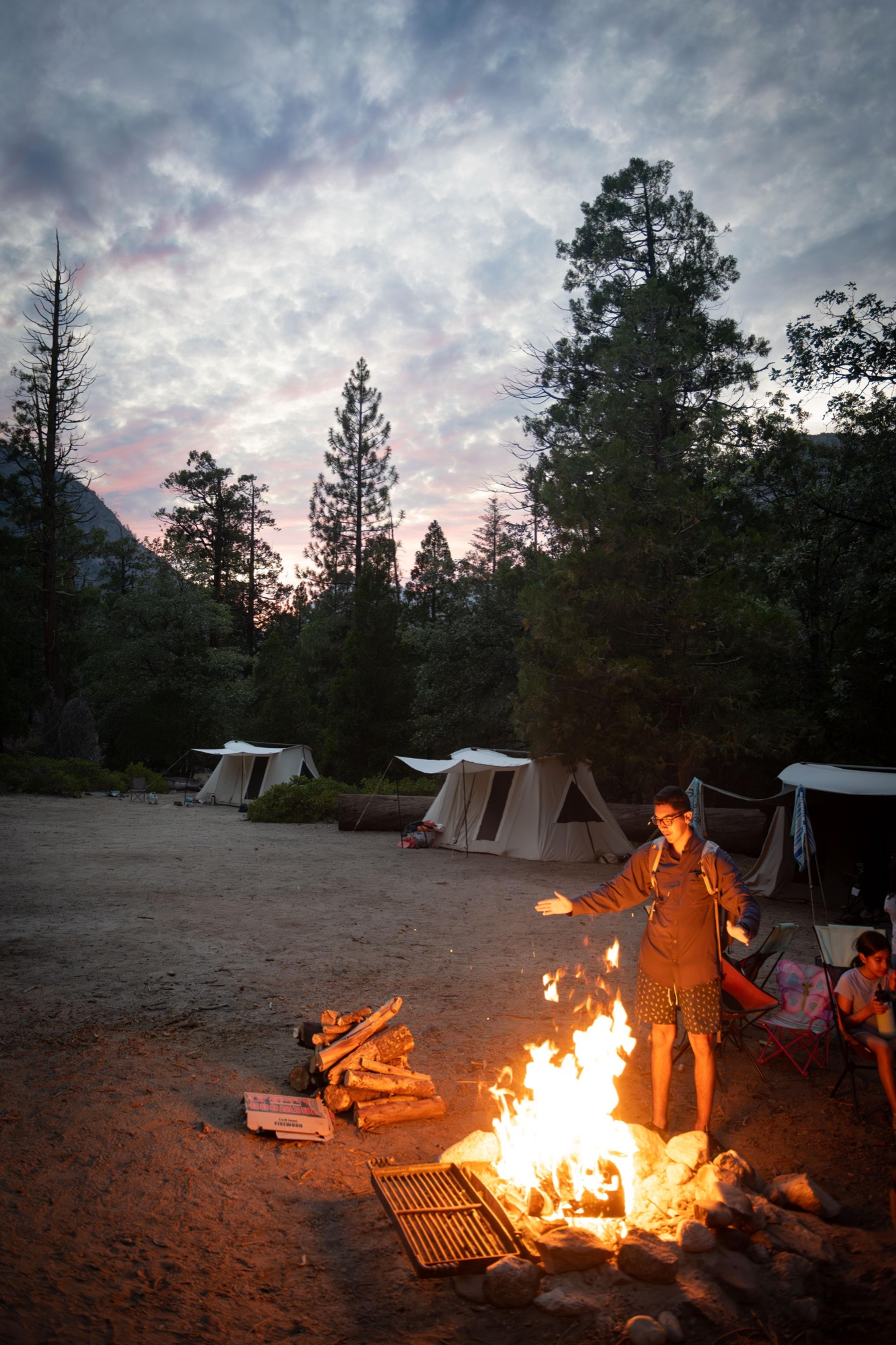 Camper tending a burning campfire at dusk in Kings Canyon with canvas tents and a pink evening sky behind Camper tending a burning campfire at dusk in Kings Canyon with canvas tents and a pink evening sky behind