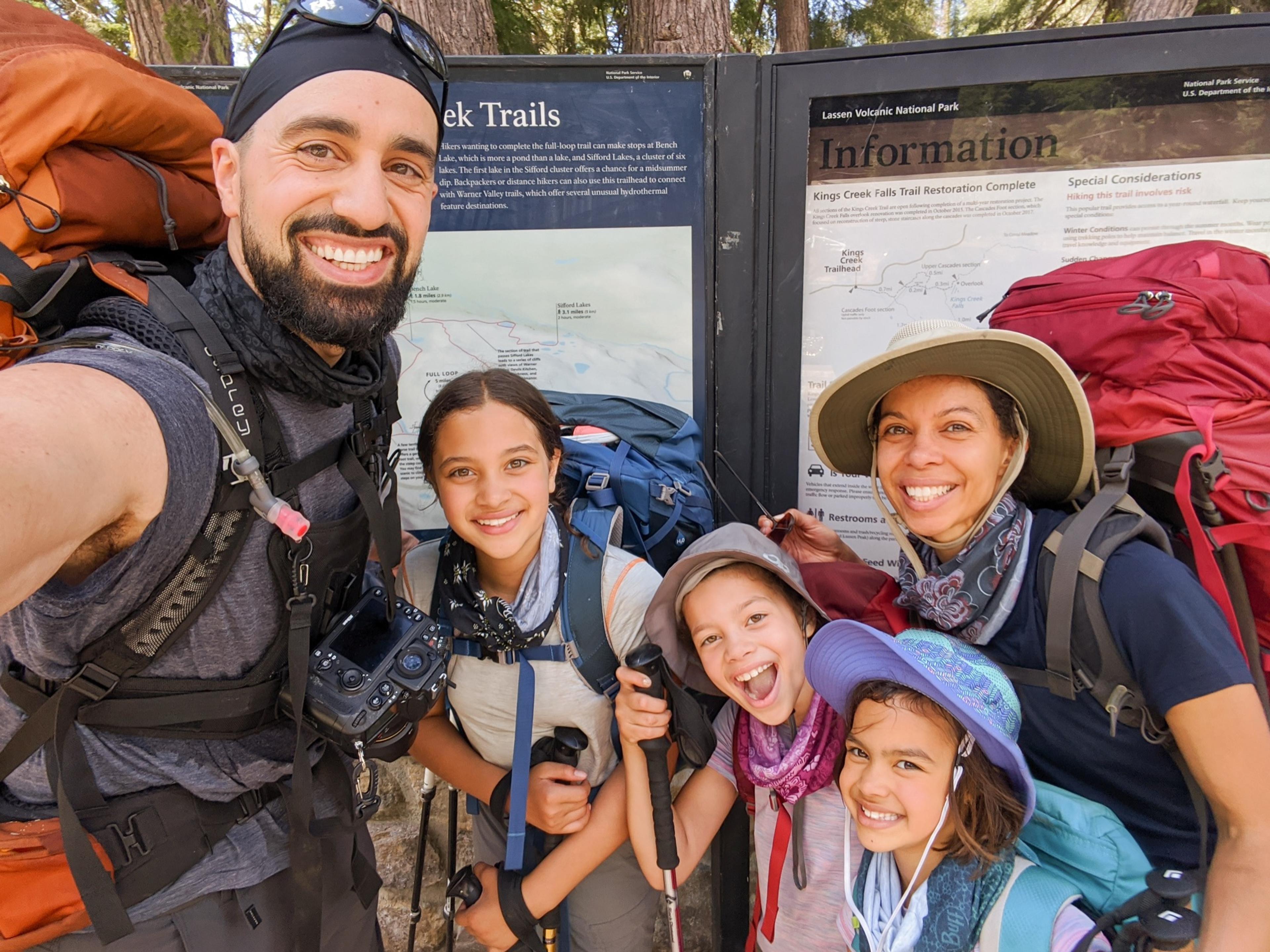 The Steele family wearing backpacking packs and smiling at the Kings Creek trailhead sign in Lassen Volcanic National Park