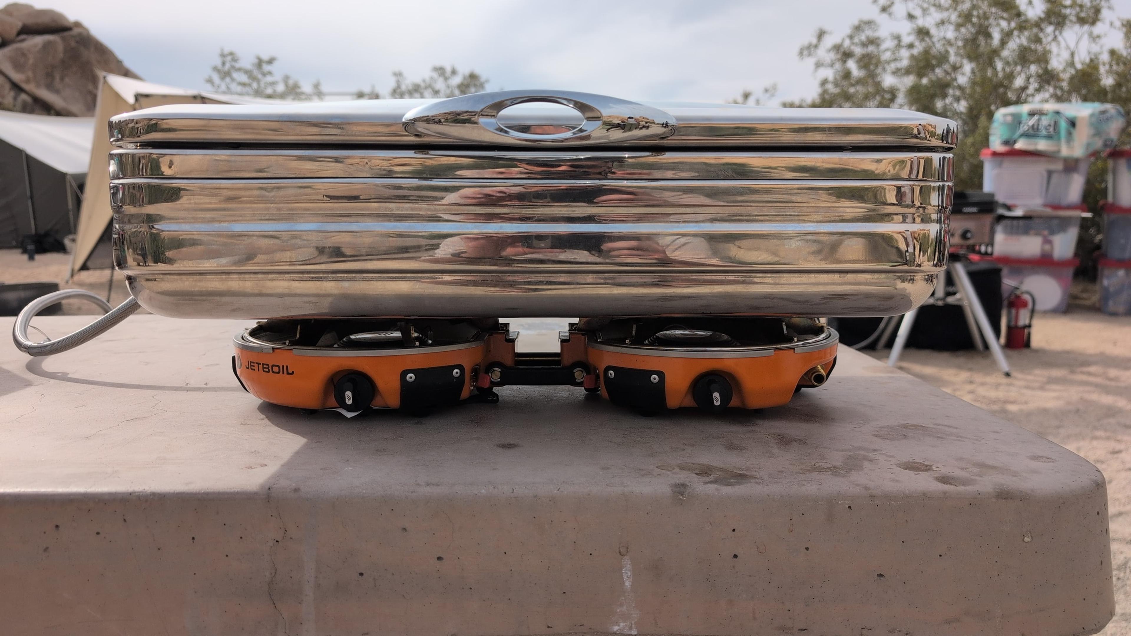 Polished stainless steel chafing dish sitting level across two orange Jetboil Genesis burners on a concrete camp table Polished stainless steel chafing dish sitting level across two orange Jetboil Genesis burners on a concrete camp table