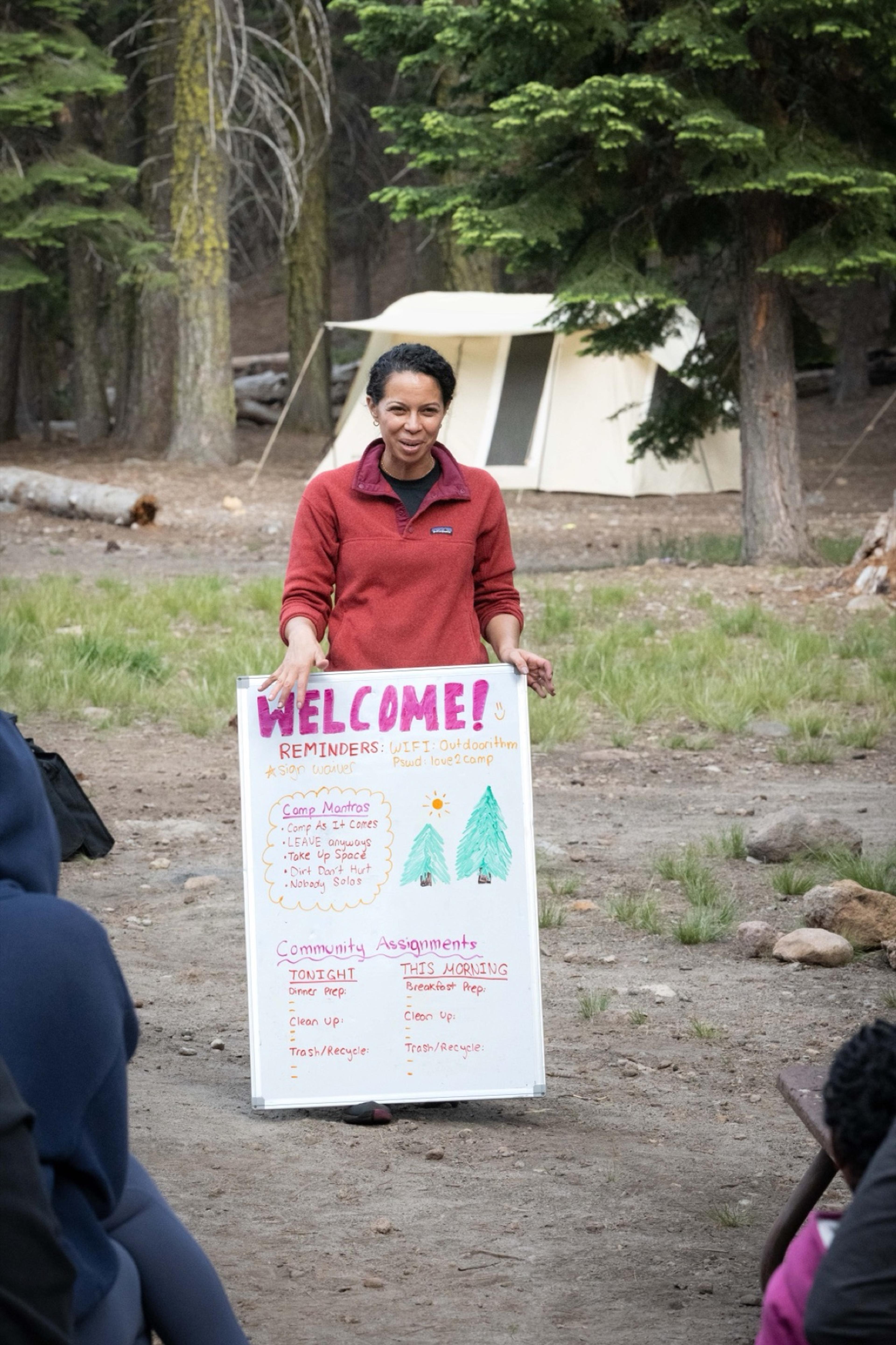 A camp host holding a hand-lettered welcome board listing camp reminders and job assignments in front of canvas tents at Lassen