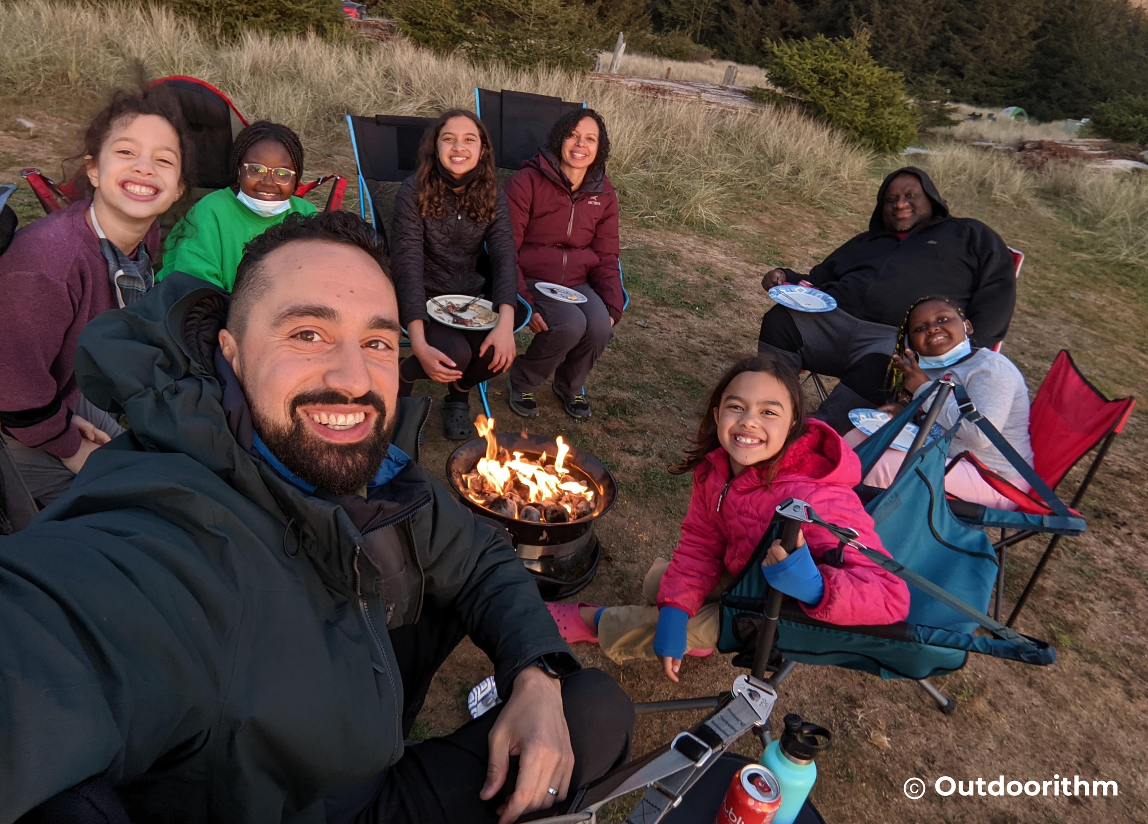 Group of campers seated around a glowing campfire at night under tall trees, finding joy and rest in the outdoors Group of campers seated around a glowing campfire at night under tall trees, finding joy and rest in the outdoors