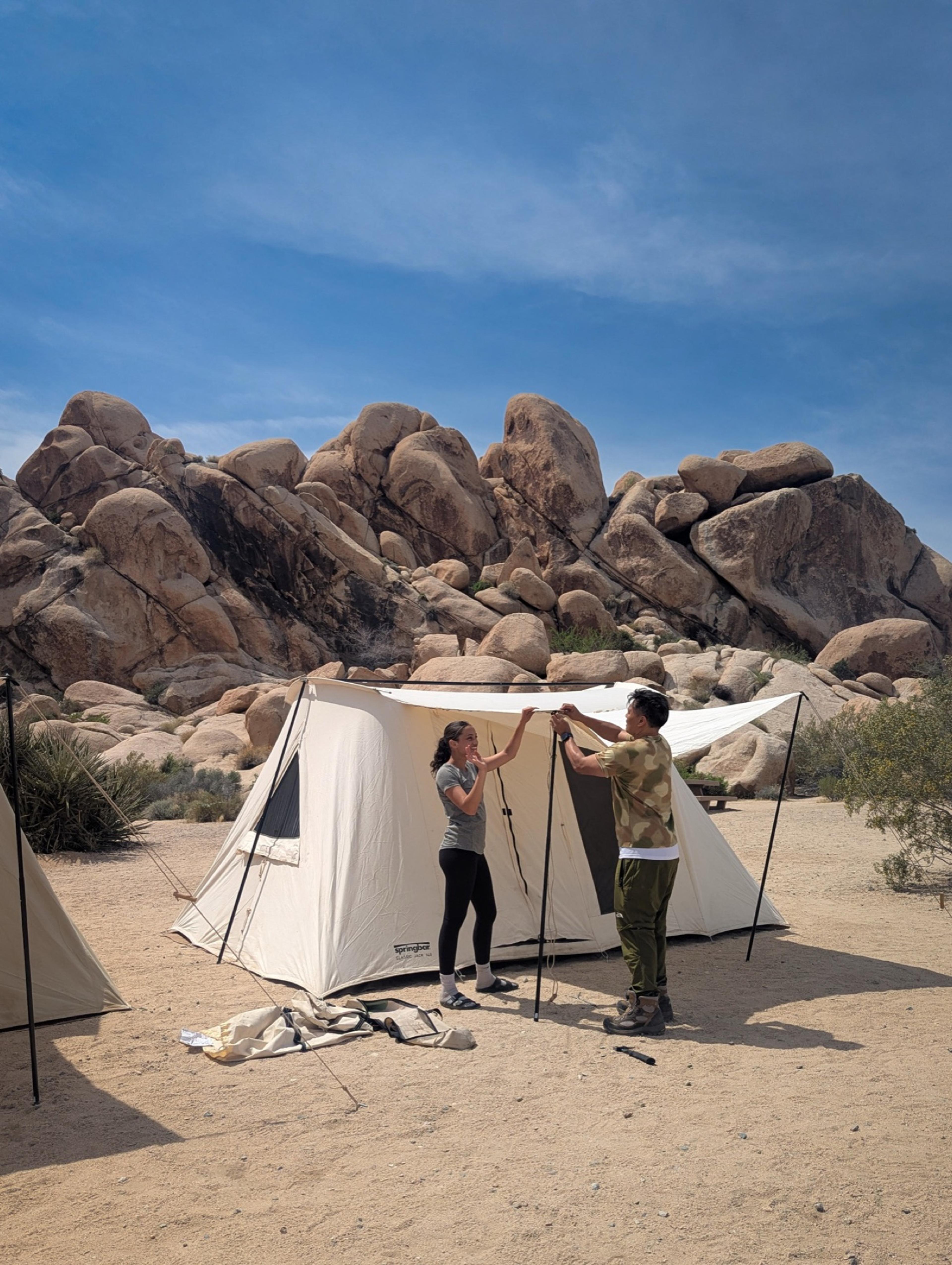 Two people raising a canvas wall tent among granite boulders at Indian Cove in Joshua Tree